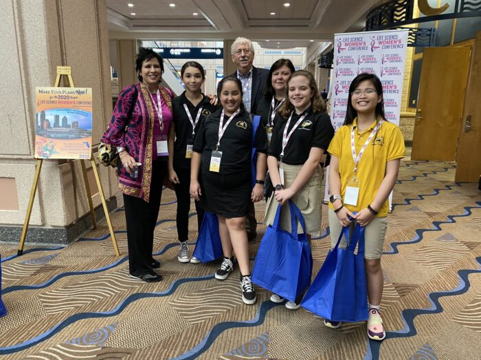 Cherie Mathews, Steve Fiske, and four young high school girls interested in STEM standing together in front of the Life Science Women's Conference 2020 sign.
