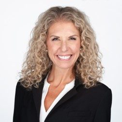 A photo of Kristy Dellisanti, smiling brightly. Her light, curly blond hair parts in the middle, cascading down to her shoulders, adorned by a black suit.