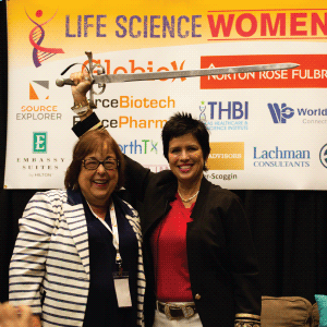 A photo of Cherie Mathews and Patti Rossman, standing proudly in front of the sponsor banner, wielding a broadsword.