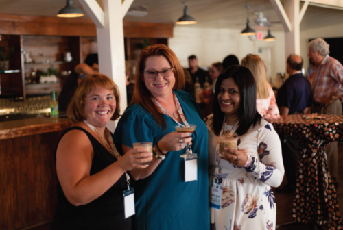 Three women standing in a row, cheering off with drinks in their hands. They're smiling and enjoying the warm atmosphere.