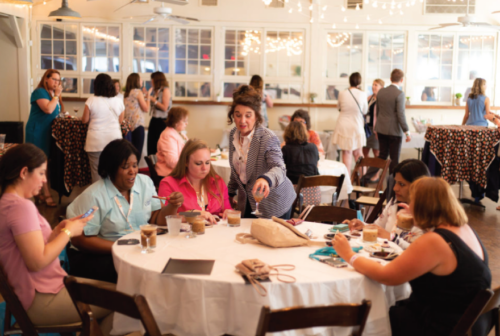 A group of conference attendees in a warm, cafe-like atmosphere, talking over food at a white-clothed table happily.