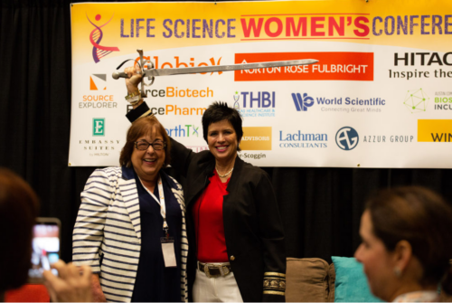 Founder Patti Rossman (left) and Cherie  Mathews (right), opening speaker, wielding a broadsword in front of a sponsor banner onstage. They're smiling, representing the power of the conference.